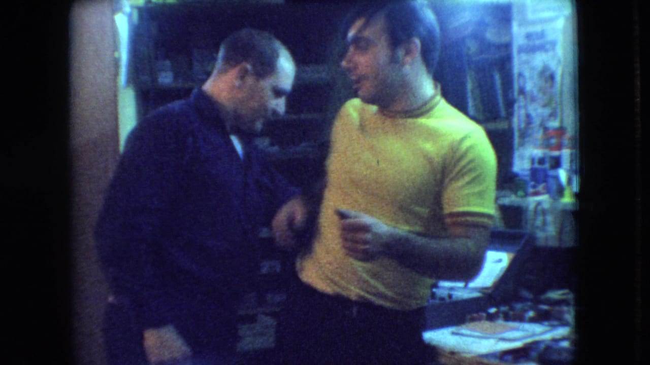 In 1966 Athens, Ohio home-movie footage, two adult men stand close together in a dim room as the man in the yellow shirt gestures toward the man in the dark sweater.