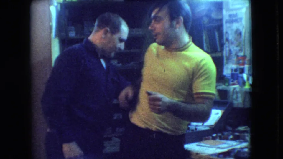 In 1966 Athens, Ohio home-movie footage, two adult men stand close together in a dim room as the man in the yellow shirt gestures toward the man in the dark sweater.