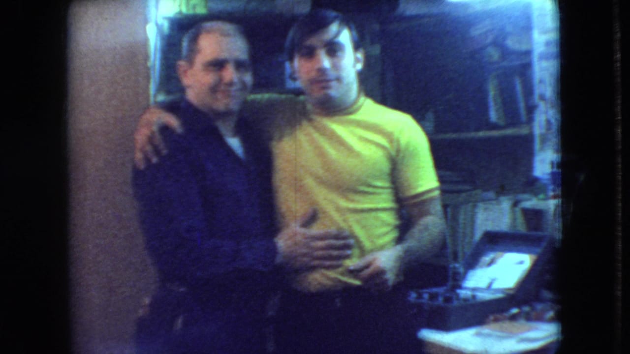 Two adult men pose with arms around each other in a small interior room.