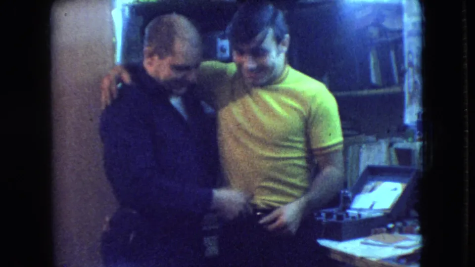 The two adult men lean together and smile in the 1966 Athens, Ohio home-movie footage, with papers and shelving visible behind them.