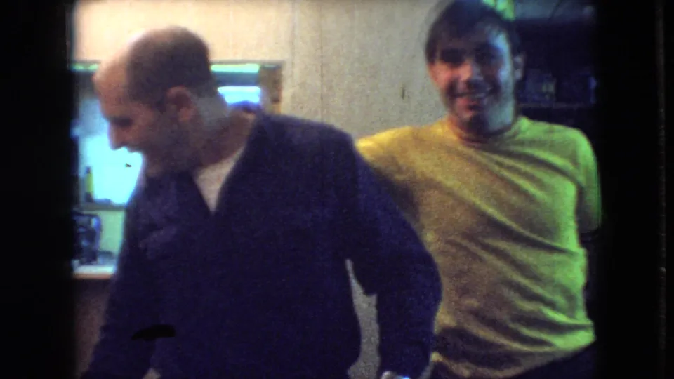 At the end of the Athens, Ohio 1966 home-movie clip, the man in the dark sweater turns or steps forward while the man in the yellow shirt smiles in the background.