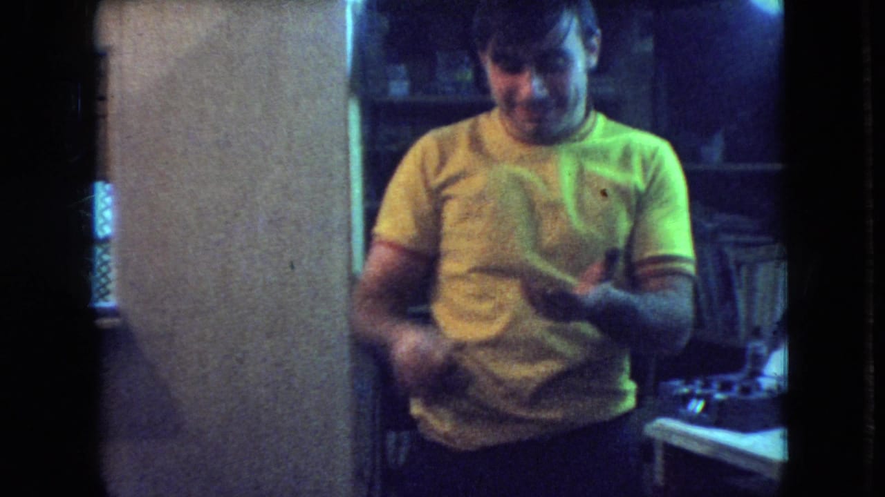 In 1966 Athens, Ohio home-movie footage, a young man in a yellow T-shirt stands inside a dim private interior, appearing to handle or inspect a small object near a workbench.