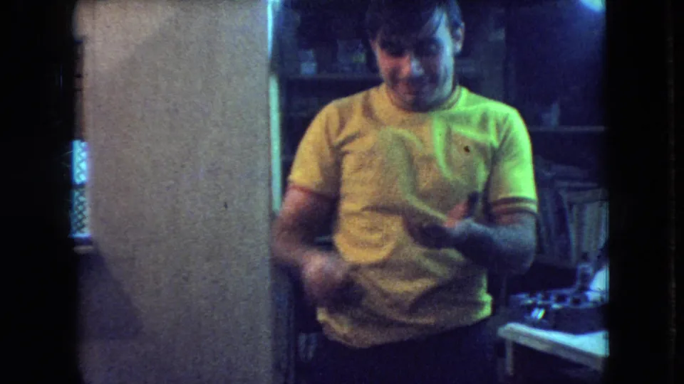 In 1966 Athens, Ohio home-movie footage, a young man in a yellow T-shirt stands inside a dim private interior, appearing to handle or inspect a small object near a workbench.