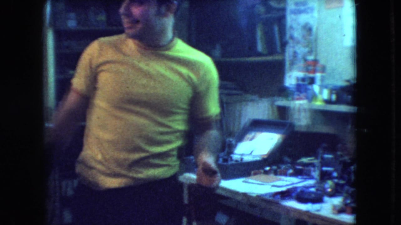 Young man stands beside a cluttered desk or workbench with a bright television screen behind him.