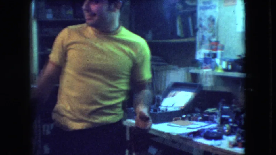 The young man stands in a dim Athens, Ohio interior beside a cluttered workbench, with a bright television screen and household items visible behind him.