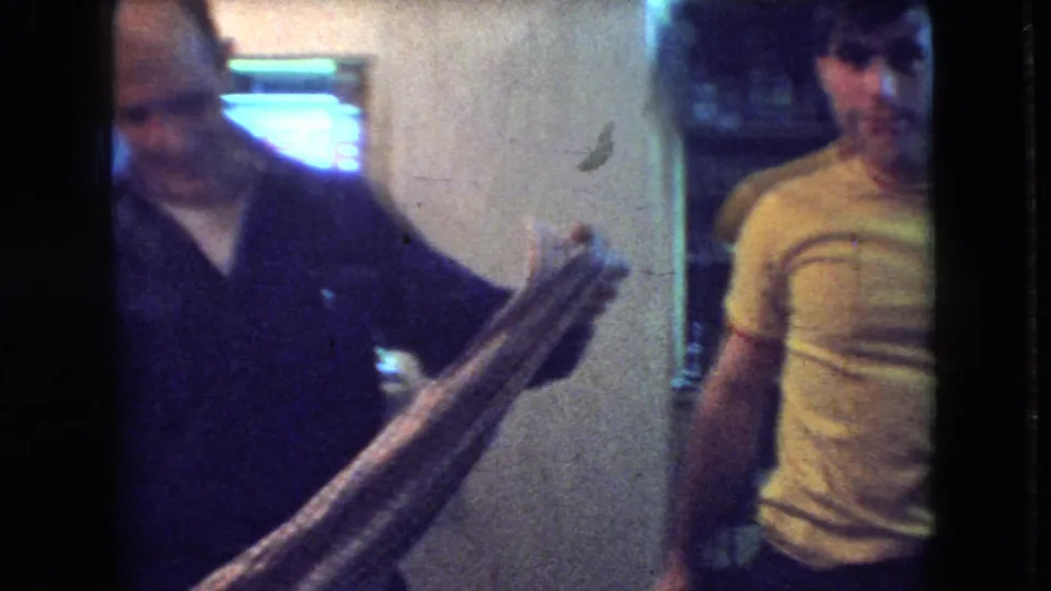An older adult in a dark shirt holds a long object in the foreground while the young man in a yellow T-shirt stands behind him in the same private Athens, Ohio interior.