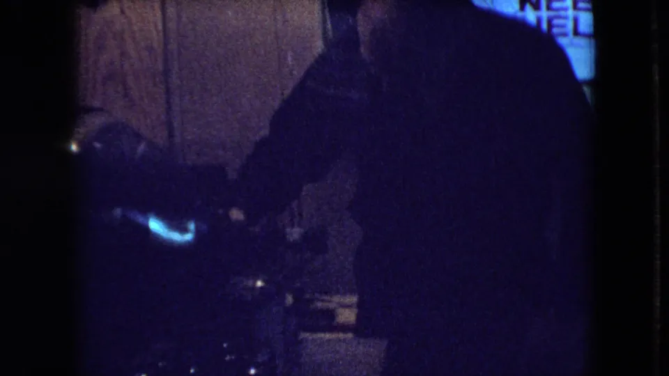 A dark, underexposed frame shows a person leaning near equipment or a work surface in the private interior, with details partly obscured by low light and film grain.