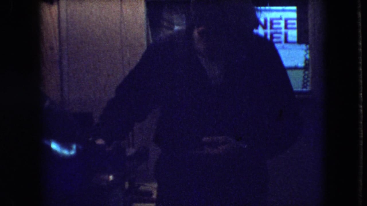 In a dim wood-paneled Athens, Ohio interior from the 1966 home-movie metadata, a man stands near a glowing television screen.