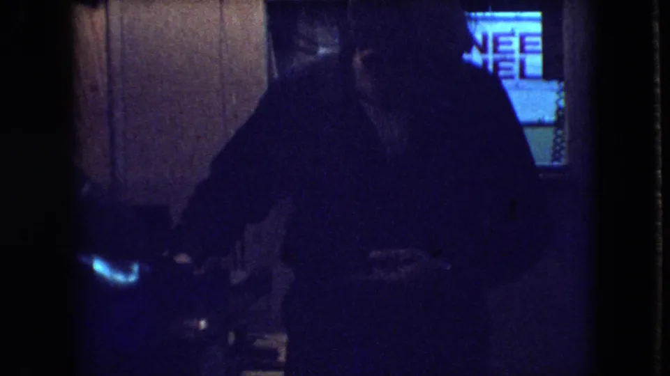 In a dim wood-paneled Athens, Ohio interior from the 1966 home-movie metadata, a man stands near a glowing television screen.