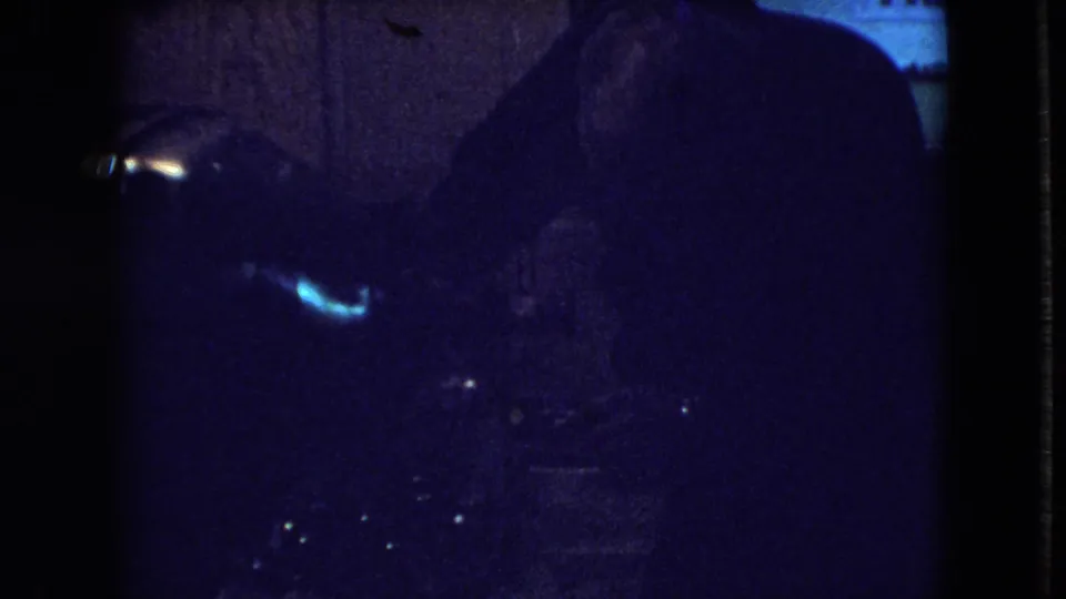The man bends forward near the television or its components, with blue light and deep shadow obscuring details in the 8mm frame.