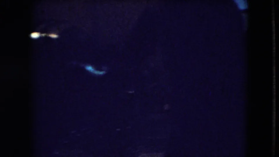 A very dark frame shows blue television light and indistinct interior shapes, likely continuing the same Athens, Ohio home-movie scene.