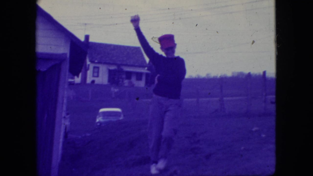 Adult man in a red cap and dark sweater raises one arm in an outdoor New Jersey residential setting in 1966, with a house and yard behind him.