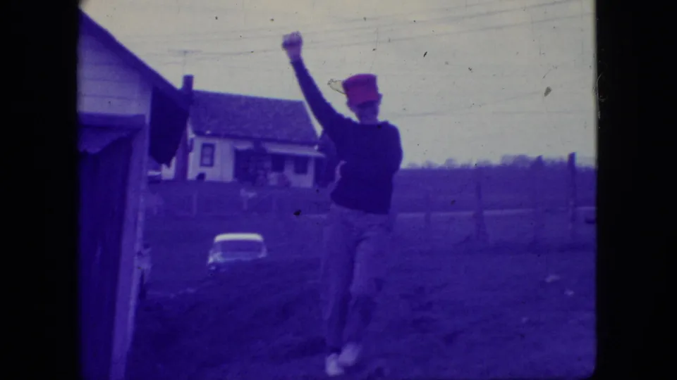 Adult man in a red cap and dark sweater raises one arm in an outdoor New Jersey residential setting in 1966, with a house and yard behind him.