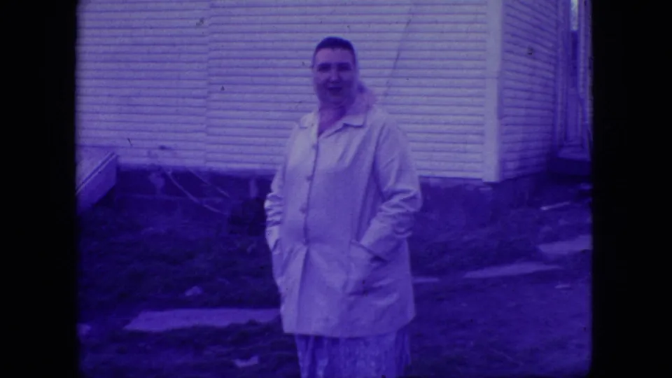 Adult woman in a light-colored jacket stands outdoors beside a residential house in the 1966 New Jersey home-movie footage.