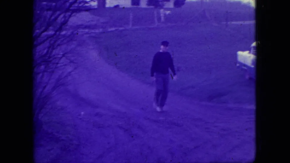 Adult man in a dark top walks away along a driveway or road in New Jersey in 1966, with a pale classic car partly visible at the edge of the frame.