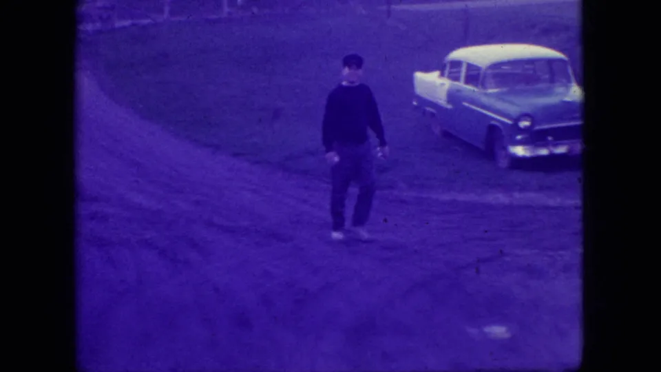 Adult man walks toward the camera near a pale 1950s-style car in the 1966 New Jersey residential outdoor scene.