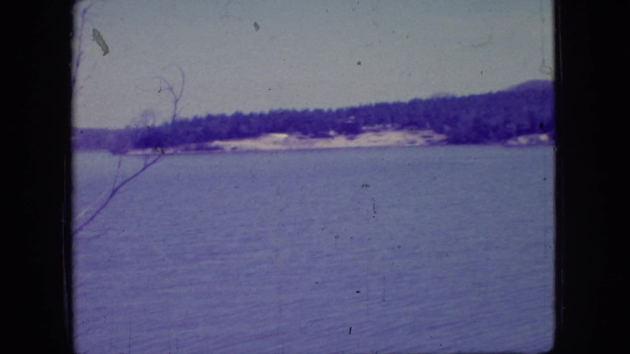 1966 New Jersey 8mm home-movie frame showing calm lake water, a distant tree-lined shoreline, low hills, pale sky, and a bare branch in the left foreground.