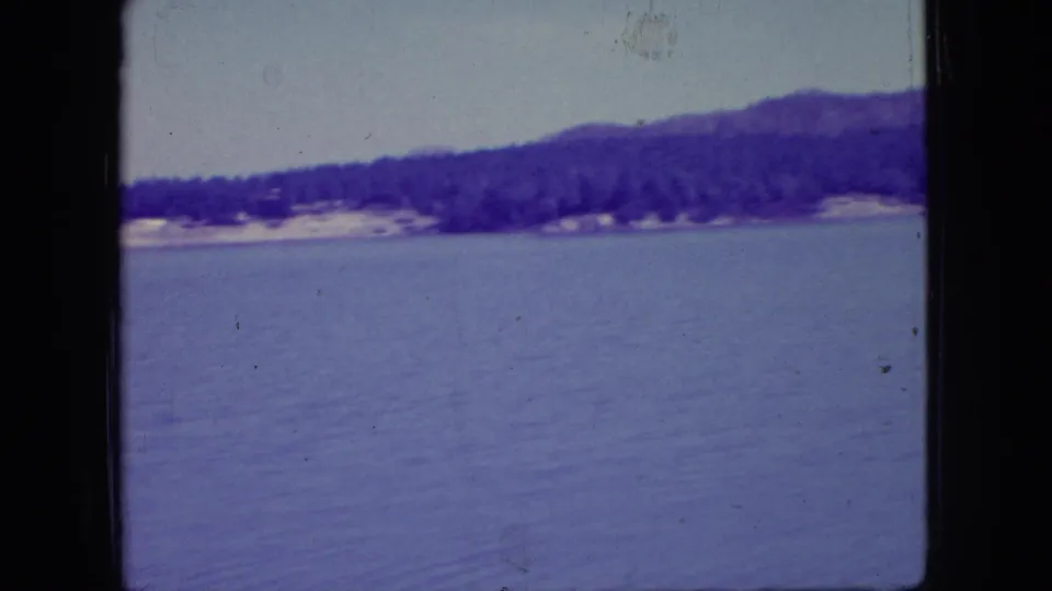 Archival 1966 New Jersey home-movie frame of a quiet lake, distant wooded bank, low hill, and soft purple-blue 8mm color shift.