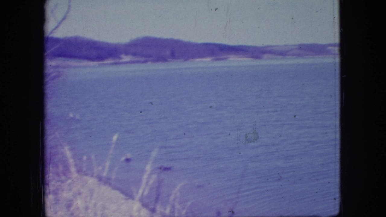 1966 New Jersey 8mm home-movie frame showing rippled lake water, distant wooded shoreline, low hills, pale sky, and visible film wear.