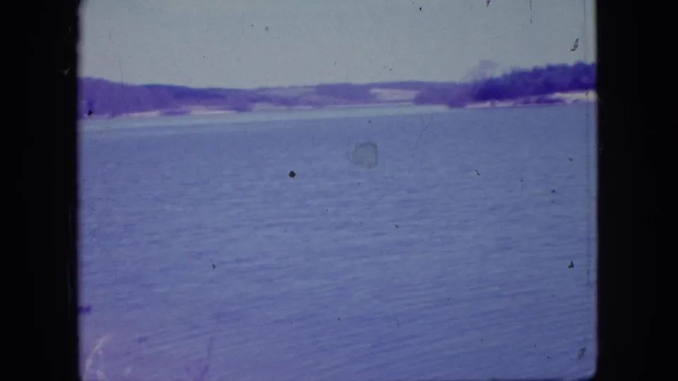 1966 New Jersey 8mm frame with open rippled water, a low forested shoreline, pale sky, purple color cast, and visible gate edges.