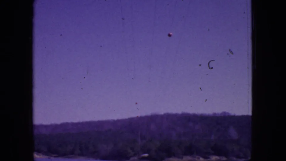1966 New Jersey 8mm frame showing a distant wooded shoreline or hillside below a pale purple sky, with dust, scratches, and dark frame borders.