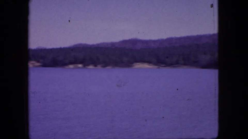 1966 New Jersey home-movie frame with lake water in the foreground, a distant wooded shore, low hills, and a pale purple-blue sky.