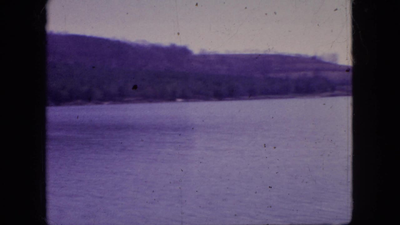 1966 New Jersey 8mm frame showing a calm lake, distant tree-lined shore, pale sky, and visible film dust along the scanned frame.