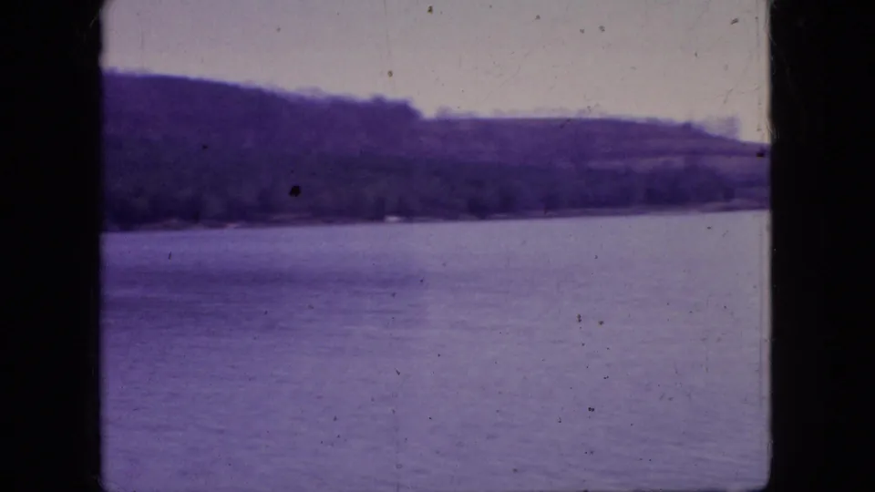 1966 New Jersey 8mm frame showing a calm lake, distant tree-lined shore, pale sky, and visible film dust along the scanned frame.