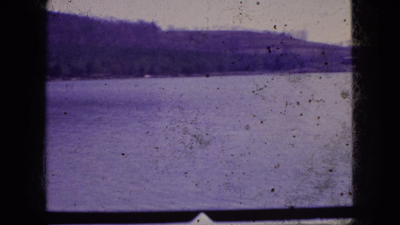 1966 New Jersey 8mm frame with a wide lake, dark wooded hillside, pale sky, dust marks, and heavy black scan borders.