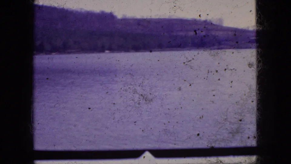 1966 New Jersey 8mm frame showing the lake and far wooded bank under a pale sky, with dust, scratches, and purple fading visible in the film scan.