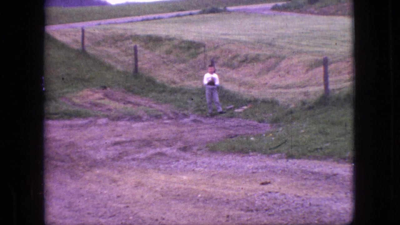 1966 New Jersey 8mm frame showing a distant person in a white shirt and light pants standing by a grassy lakeside field, fence posts, dirt track, and open farmland.