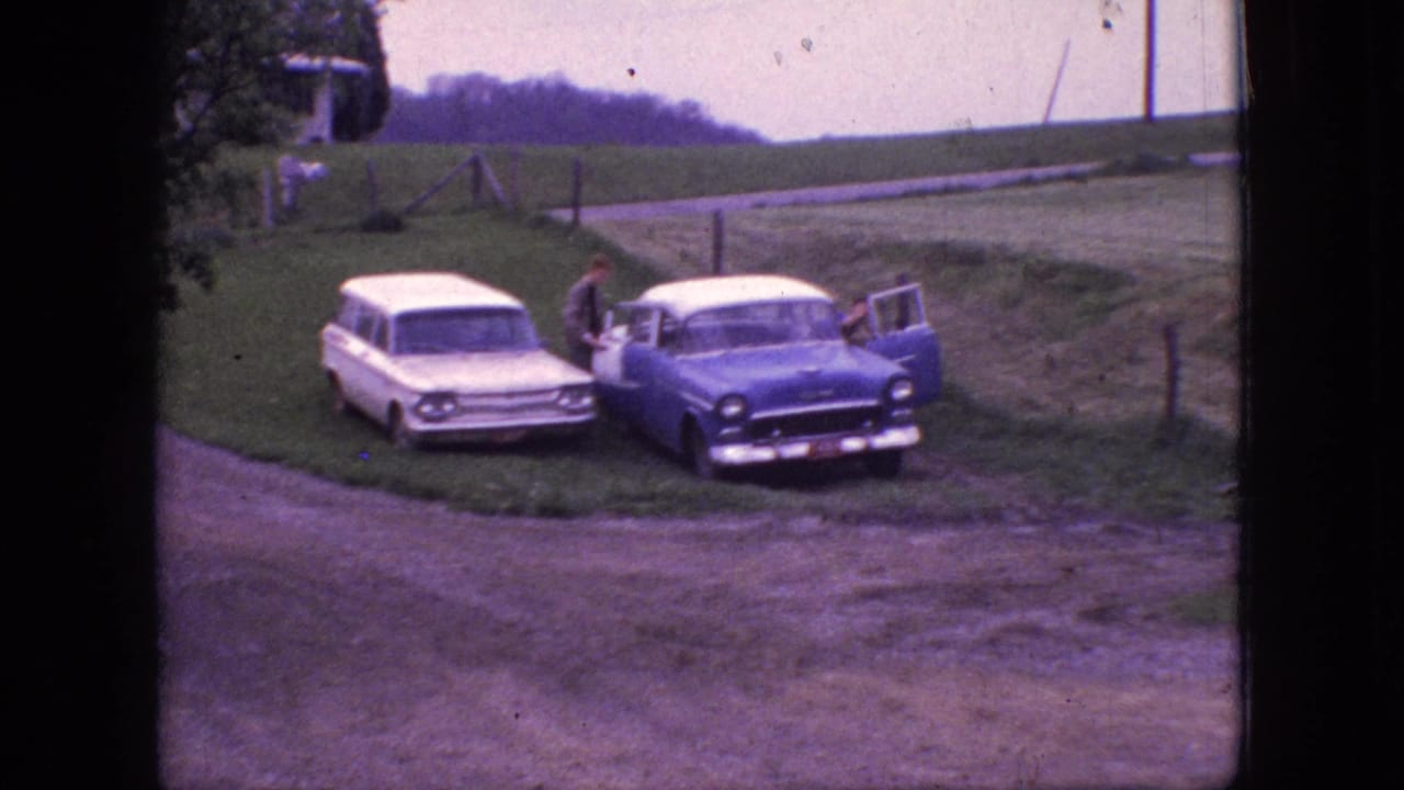 A man stands beside the blue sedan as its passenger-side door is open near a white station wagon.