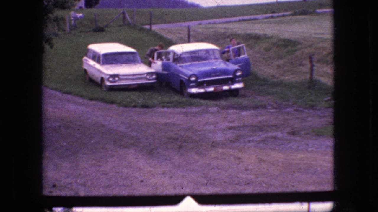 1966 New Jersey home-movie frame with the blue-and-white sedan and white station wagon parked beside a rural dirt road, framed by grass, fence posts, and open fields.