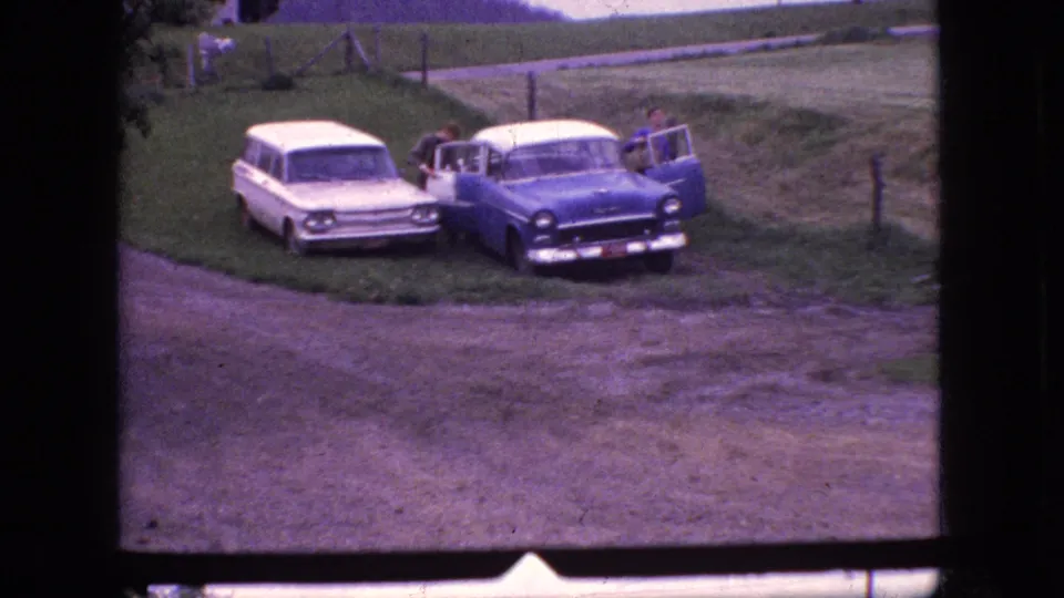 1966 New Jersey home-movie frame with the blue-and-white sedan and white station wagon parked beside a rural dirt road, framed by grass, fence posts, and open fields.