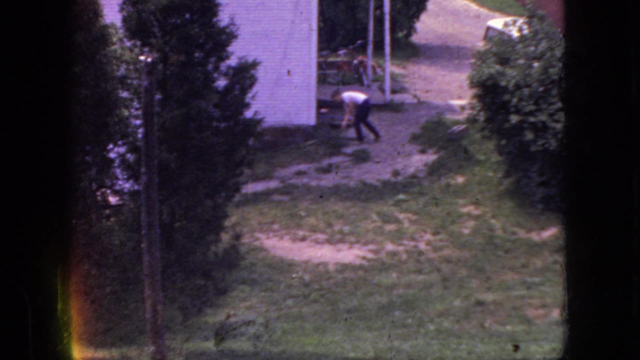 1966 New Jersey home-movie frame showing a man in a white shirt and dark pants bending or working in a grassy yard near a white building and trees.