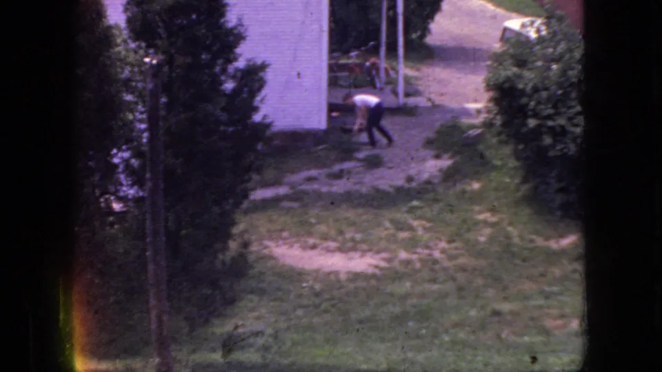 1966 New Jersey home-movie frame showing a man in a white shirt and dark pants bending or working in a grassy yard near a white building and trees.