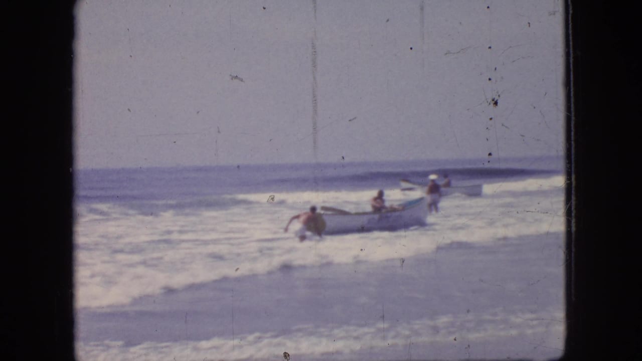 1966 New Jersey beach frame showing several people in shallow ocean water around a small boat as waves break near the shore.