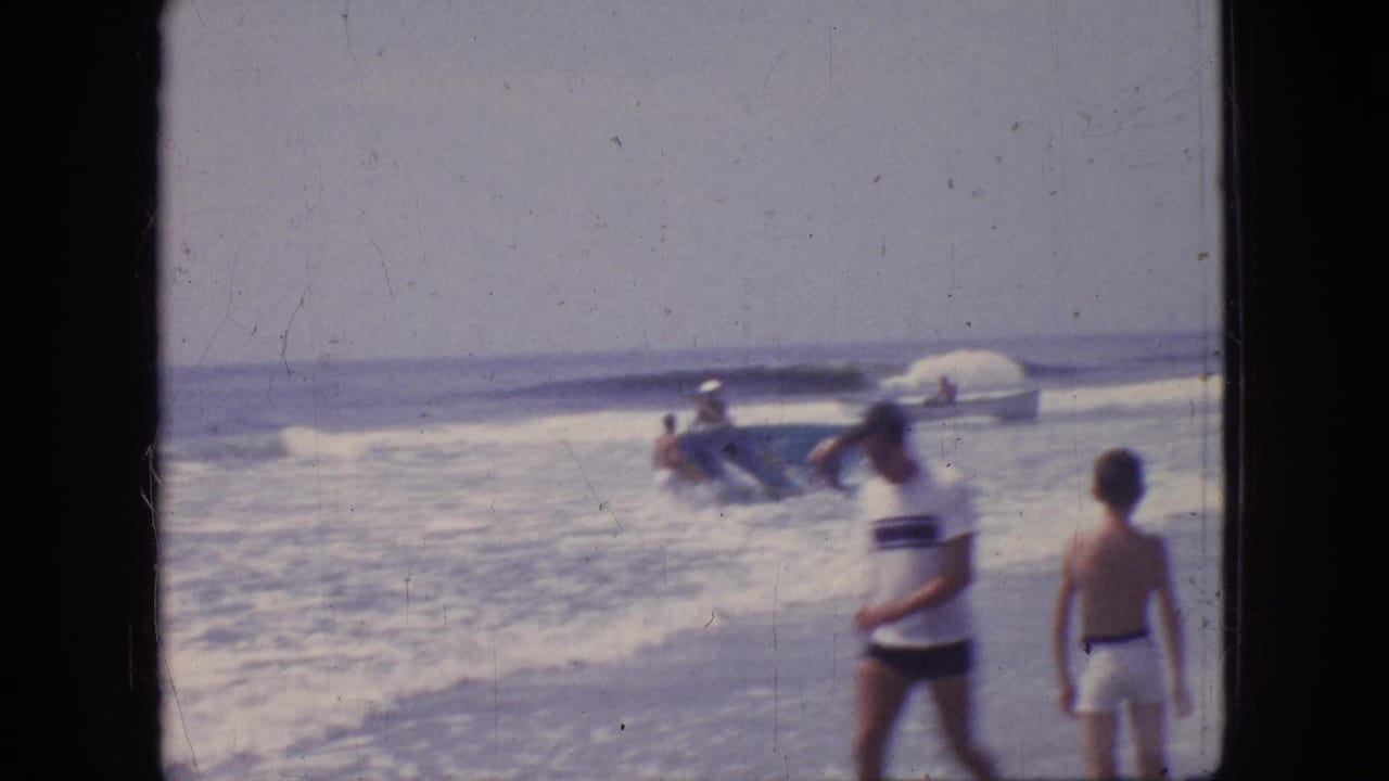 1966 New Jersey beach frame with two figures near the foreground and several people around a small boat in the breaking surf.