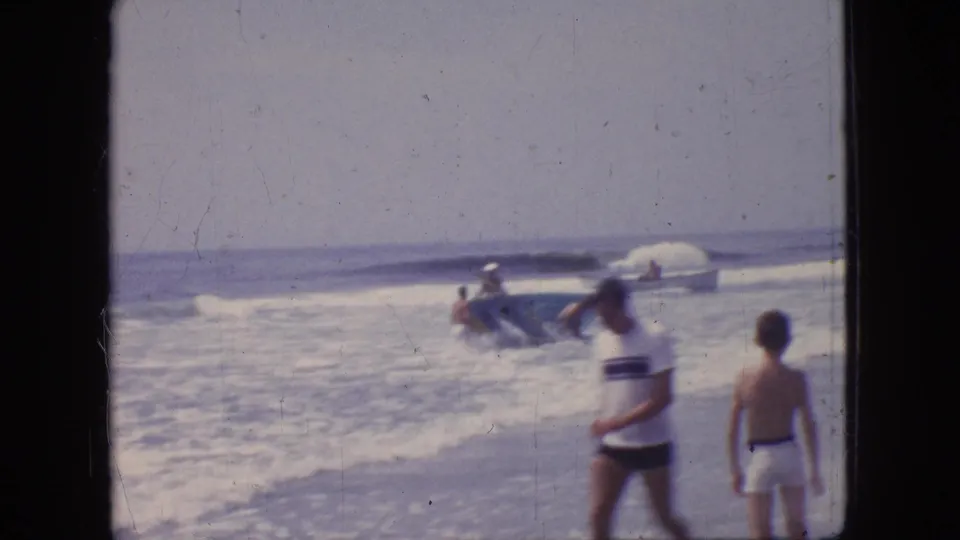 1966 New Jersey beach frame with two figures near the foreground and several people around a small boat in the breaking surf.