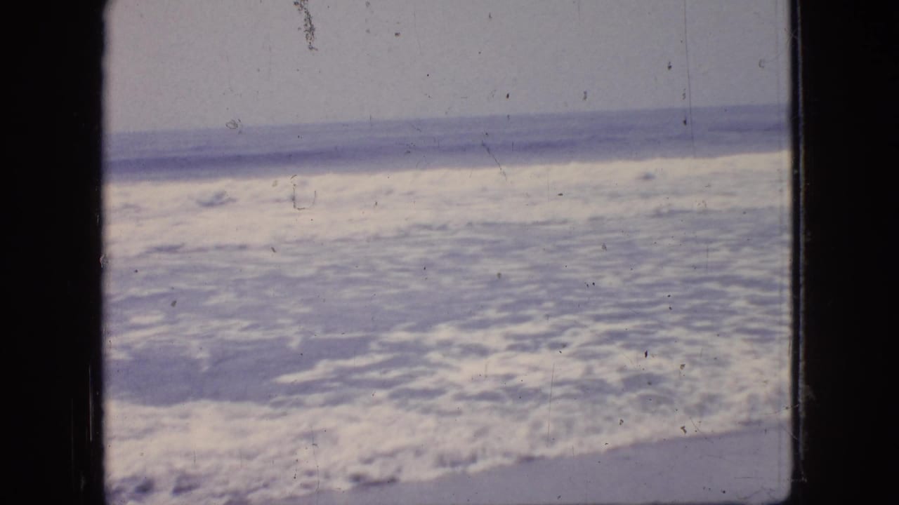 1966 New Jersey home-movie frame showing churning ocean surf viewed through a dark window-like frame, with soft focus and visible film wear.