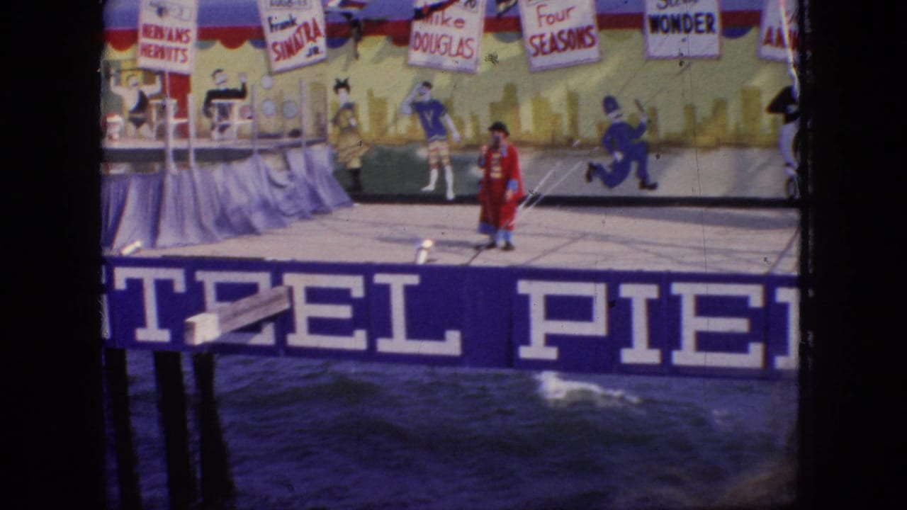 1966 New Jersey 8mm frame showing a red-coated clown or ringmaster on a Steel Pier stage above choppy water, with painted backdrop figures and promotional signs overhead.