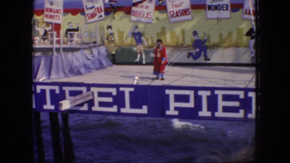 1966 New Jersey 8mm frame showing a red-coated clown or ringmaster on a Steel Pier stage above choppy water, with painted backdrop figures and promotional signs overhead.