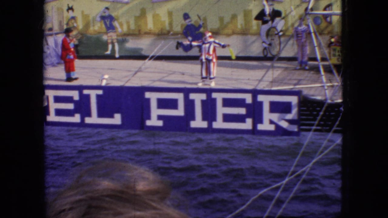 1966 New Jersey 8mm frame showing a striped clown performing on the Steel Pier stage beside a red-coated performer, with water, rigging, and painted scenery visible.