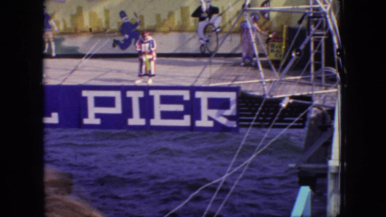 1966 New Jersey home-movie frame of a striped clown on the Steel Pier stage near visible rigging, with painted sports figures behind and choppy water below.