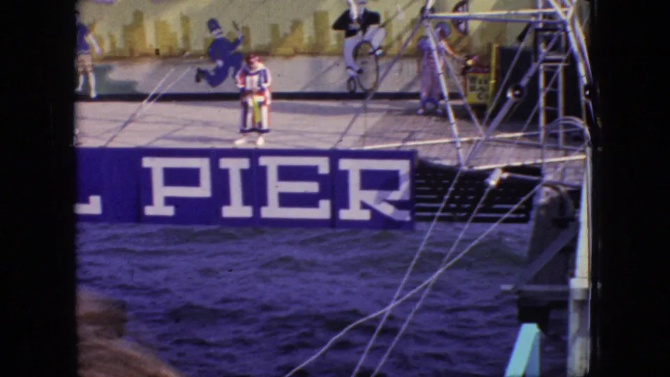 1966 New Jersey home-movie frame of a striped clown on the Steel Pier stage near visible rigging, with painted sports figures behind and choppy water below.