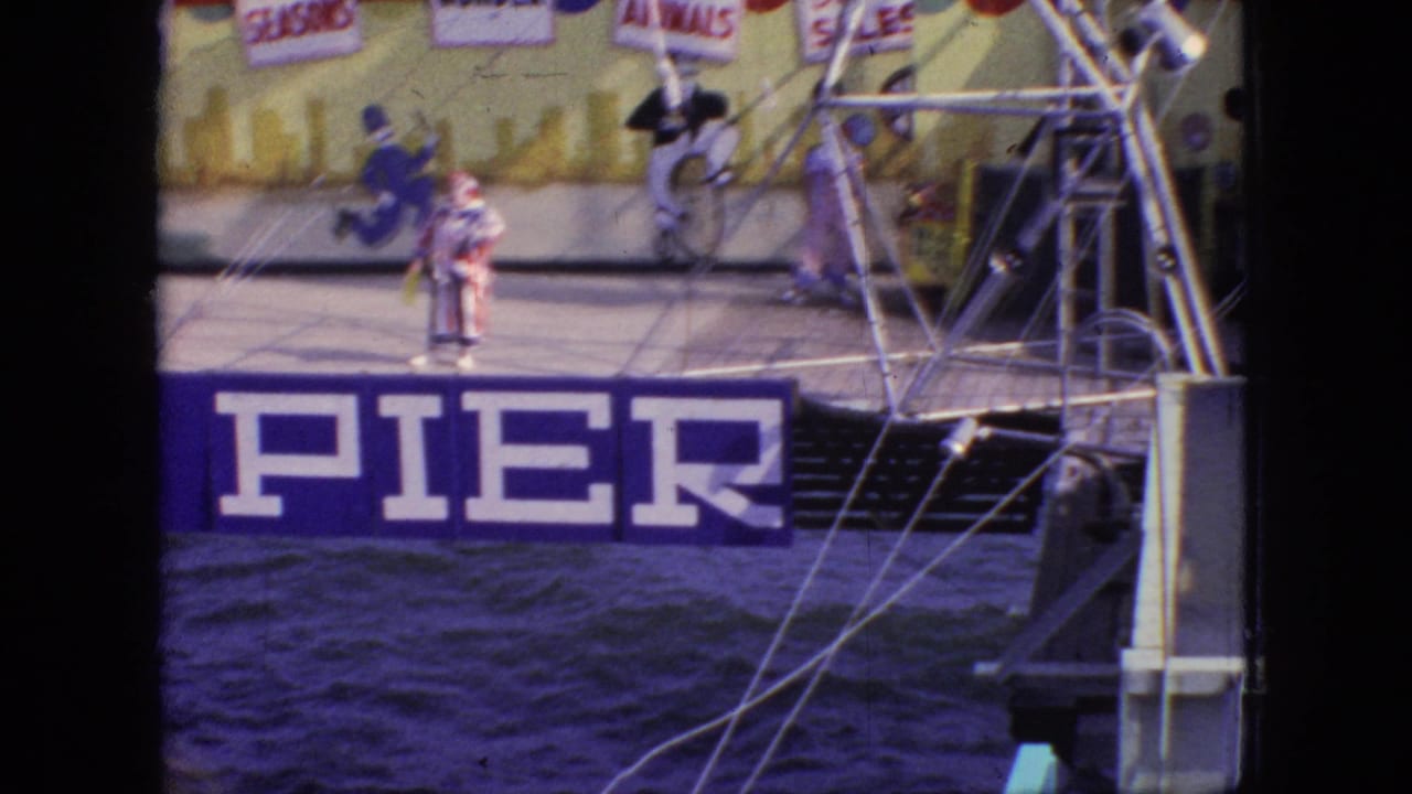 1966 New Jersey 8mm frame showing clown performers on a pier stage above dark water, with metal pier structure and a large blue PIER banner in view.