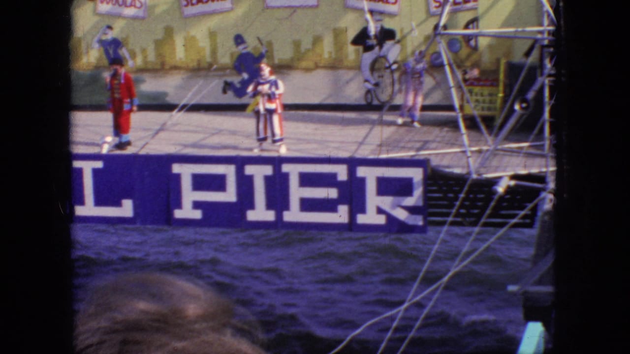 1966 New Jersey 8mm frame with a red-coated performer and striped clowns on a pier stage, backed by circus-style signage and metal structure.
