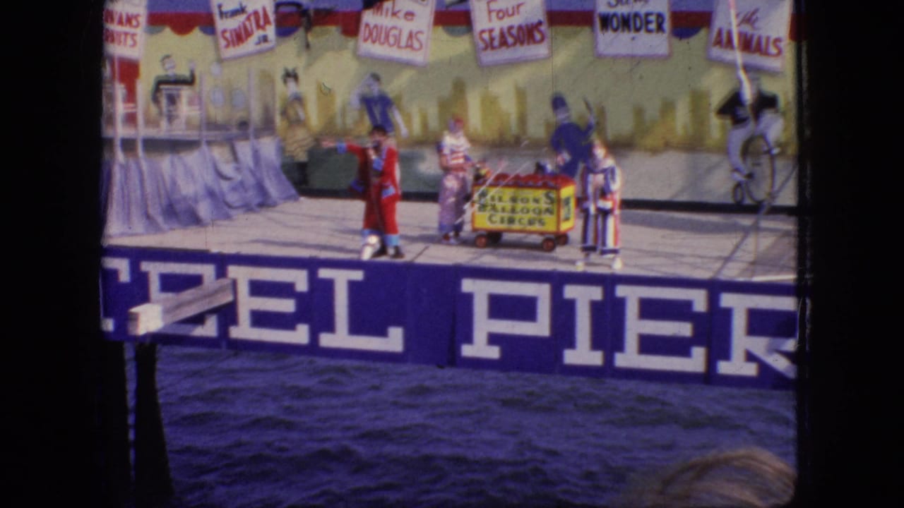 1966 New Jersey 8mm frame showing clowns and a red-coated performer beside a small circus cart on a pier stage with performer-name placards overhead.