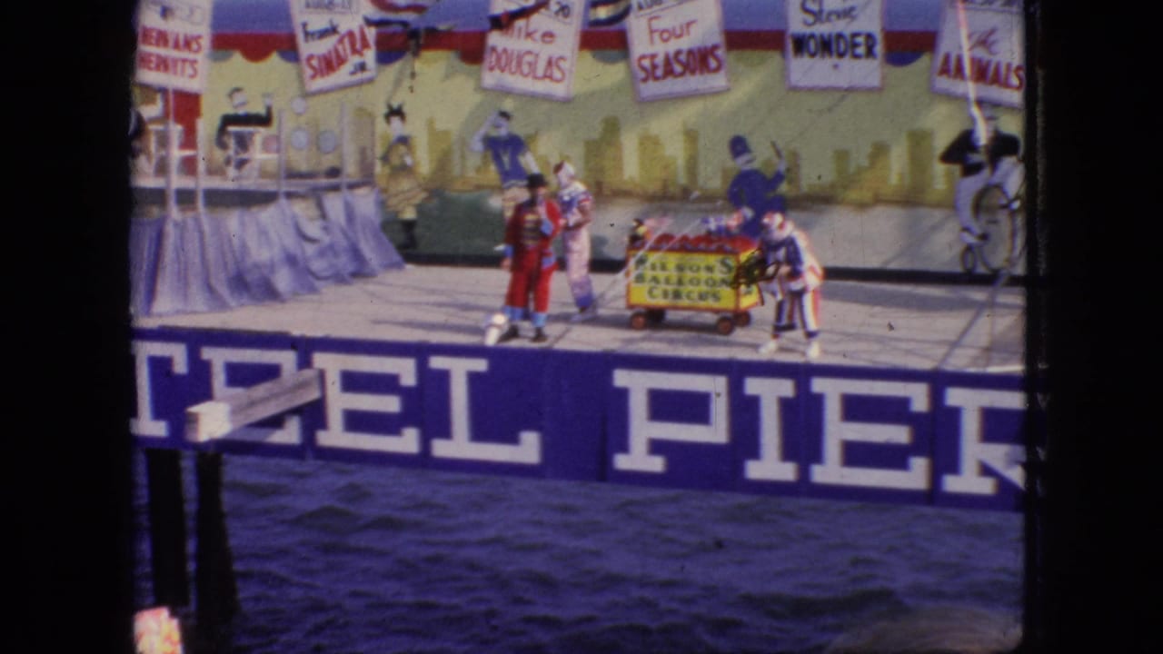 1966 New Jersey 8mm frame of the pier circus act with performers grouped around a small cart, the blue PIER banner, water, and overhead performer-name signs visible.