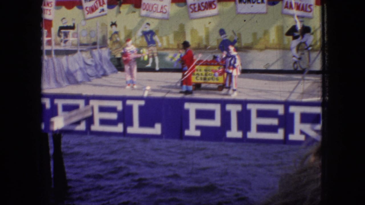 1966 New Jersey home-movie view of performers in bright clown costumes on a Steel Pier stage, filmed from across the water with a painted backdrop behind them.